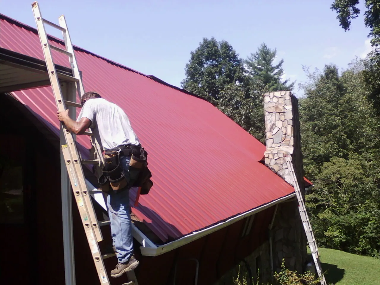 Roofer on a ladder beside a red metal roof and stone chimney — Soffit & Fascia Repair by Roofing Brothers in Michigan City